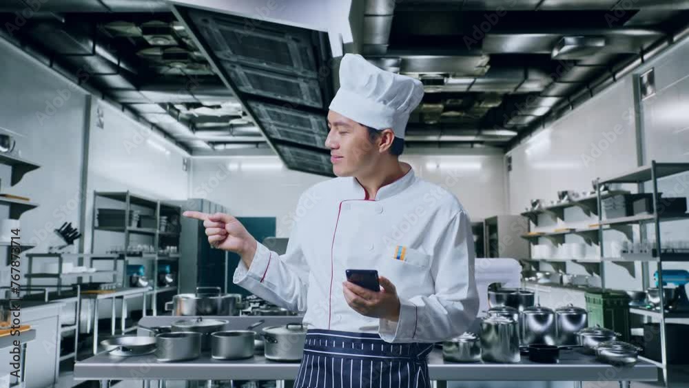 Asian Man Chef Using Smartphone And Pointing To Side While Standing In Kitchen Asian Man Chef Using Smartphone And Pointing To Side While Standing In Kitchen