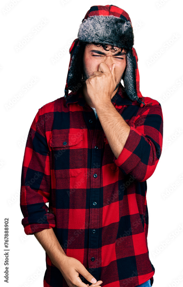 Hispanic young man with beard wearing fluffy earmuff hat smelling something stinky and disgusting, intolerable smell, holding breath with fingers on nose. bad smell Hispanic young man with beard wearing fluffy earmuff hat smelling something stinky and disgusting, intolerable smell, holding breath with fingers on nose. bad smell