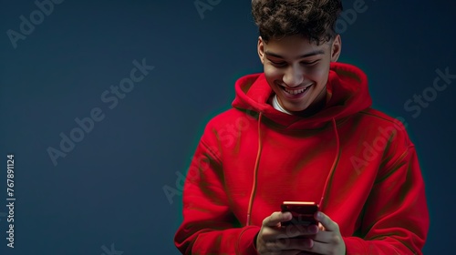 young man in a red sweatshirt smiling, using a phone on a dark blue background, professional color correction.