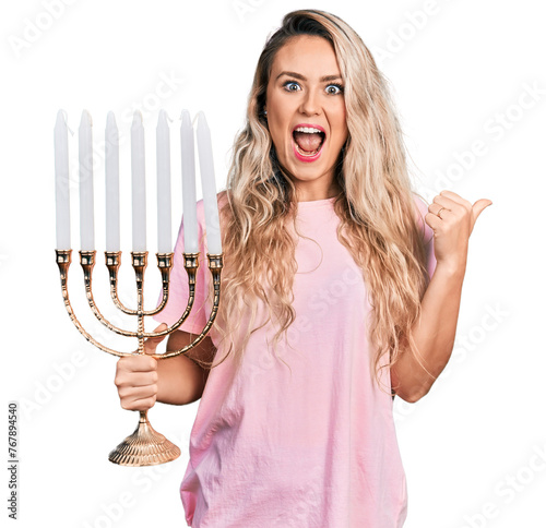 Young blonde woman holding menorah hanukkah jewish candle pointing thumb up to the side smiling happy with open mouth photo