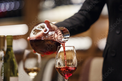 Professional female sommelier pours red wine from decanter to the glass, close up image. Woman waiter pouring alcoholic drink being in cellar of wine shop. photo