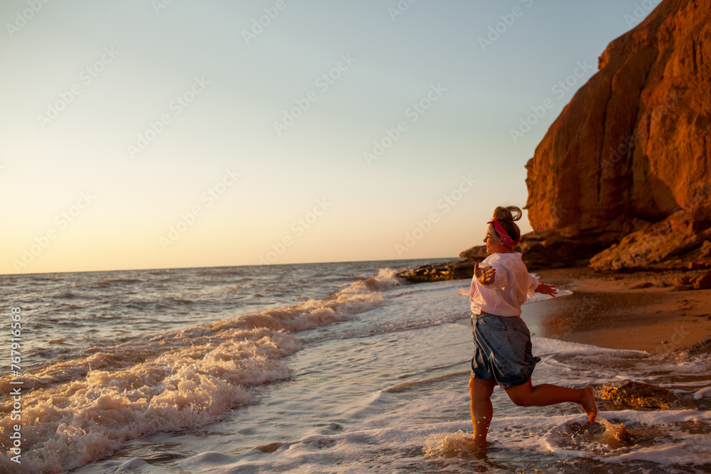 A beautiful plus size woman dressed solid  pink swimsuit, jeans shorts, sunglasses and white shirt walks along the seashore against the sand beach and sea or ocean during the sunset
