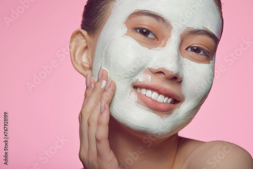 Close up portrait of a young female asian model applying clay face mask on a pink isolated.