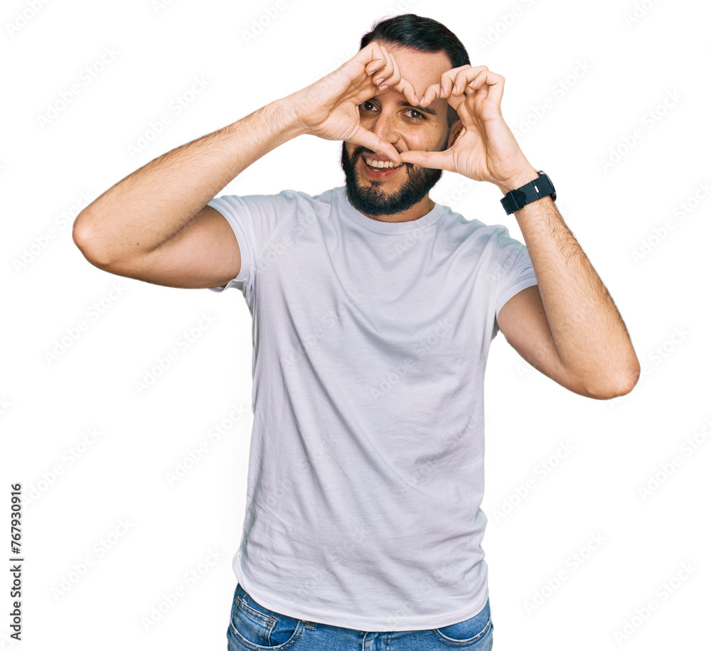 Young man with beard wearing casual white t shirt doing heart shape with hand and fingers smiling looking through sign Young man with beard wearing casual white t shirt doing heart shape with hand and fingers smiling looking through sign