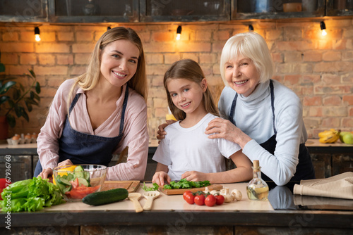 Smiling grandmother, mother and daughter doing some healthy organic food in kitchen with look at camera. Happy granny spending time with family in retired, cooking dinner together.