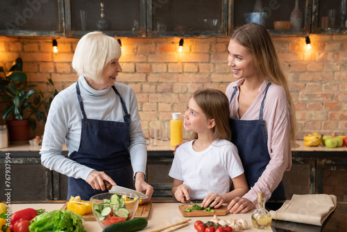Cheerful family cooking vegetarian healthy breakfast in kitchen. Grandmother with little girl and mother help prepare salad for dinner, sharing a family secret recipe