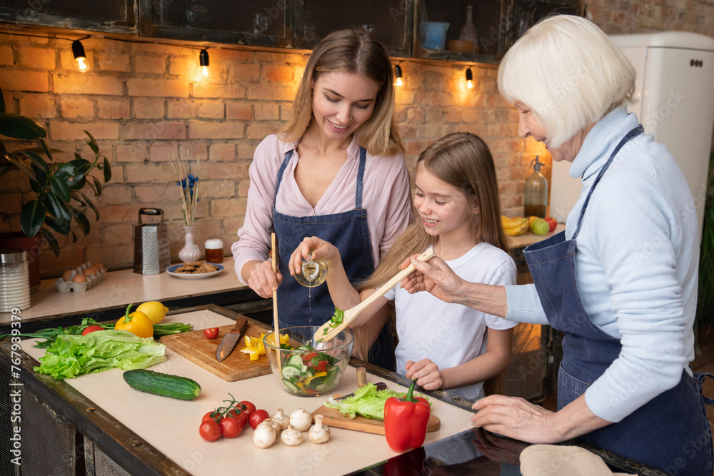 Little cute girl pours oil in fresh salad while young beautiful mother and grey hair grandmother helping her to mix it by wooden spoons in home kitchen. Fresh vegetables and salad on desk