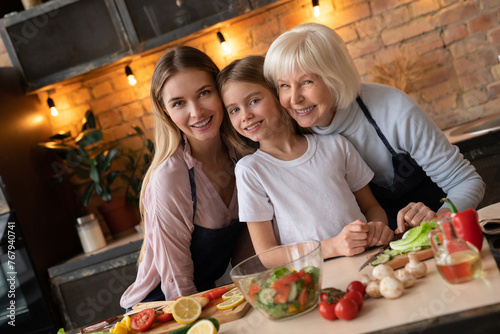 Happy family smiling and looking in camera while preparing healthy salad in kitchen. Vegetables and fruits on table. Grandmother, mother, daughter spend time together in kitchen