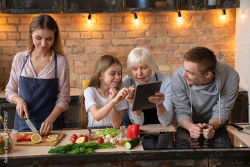 Grandmother showing something to her granddaughter in the tablet in kitchen while mother preparing fresh organic vegetables for vegetarian salad. Happy farther looking on his family photo