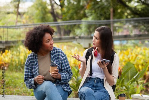 Two women sitting on a bench in a park, one of them holding a cup. They are talking to each other