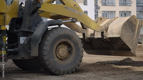A construction site is being cleared by a yellow bulldozer with strong tires, moving dirt and debris. The vehicles wheels are covered in mud and tread marks are visible on the asphalt