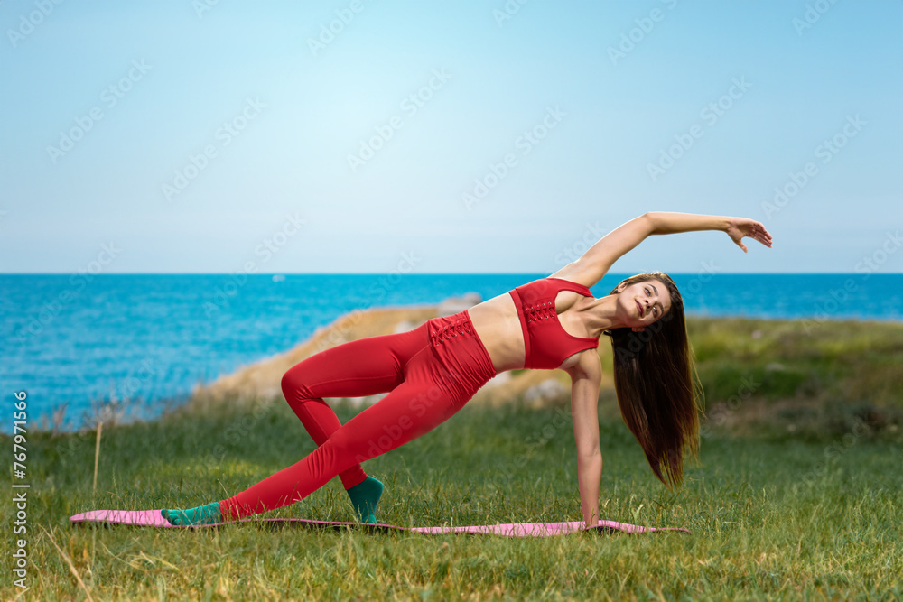 Fototapeta premium Pretty healthy woman doing active training exercise on a yoga mat near the ocean. Coastal yoga tranquility: A young woman stretches alone, connecting with the soothing ocean