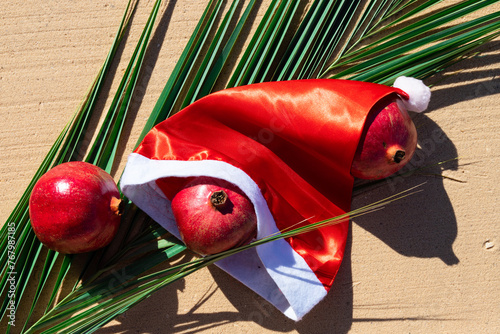 Hat of Santa Claus on the background of a palm leaf, with pomegranate fruits on the sandy beach of the Red Sea. Tropical Christmas card. photo