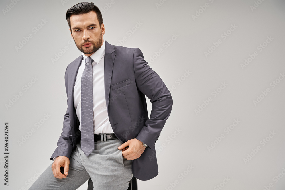 Fototapeta premium handsome and confident businessman in elegant formal wear sitting on high stool on grey background