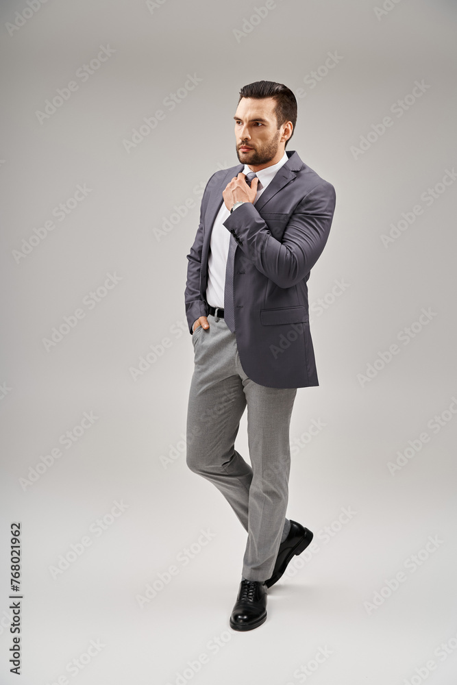 handsome man in sharp and stylish suit adjusting his tie and posing with hand in pocket on grey handsome man in sharp and stylish suit adjusting his tie and posing with hand in pocket on grey