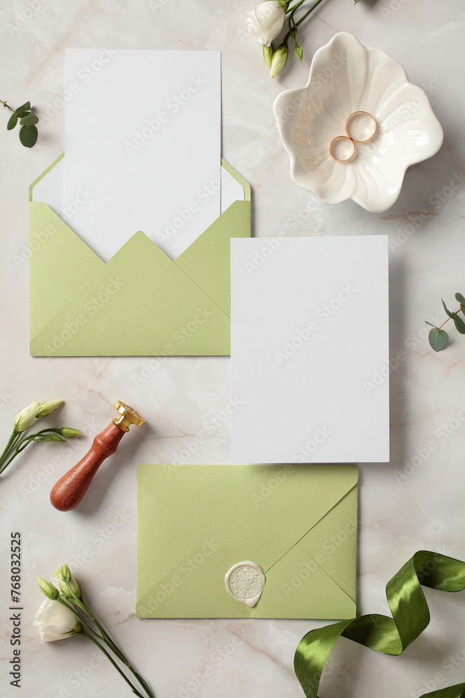 Wedding invitation cards mockups with olive envelopes, gold rings, flowers, wax seal stamp on stone background. Flat lay, top view, copy space