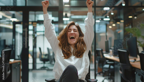 Young Woman Startup Founder Expressing Joy and Excitement, Making a Winning Gesture with Arms Raised While Sitting on Chair in Office