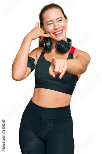 Beautiful blonde woman wearing gym clothes and using headphones smiling doing talking on the telephone gesture and pointing to you. call me.
