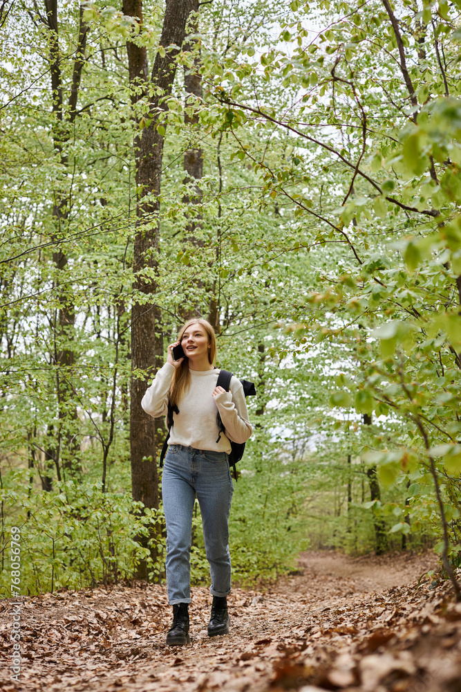 Pretty blonde woman traveler with backpack talking by phone walking in forest scenery - obrazy, fototapety, plakaty 