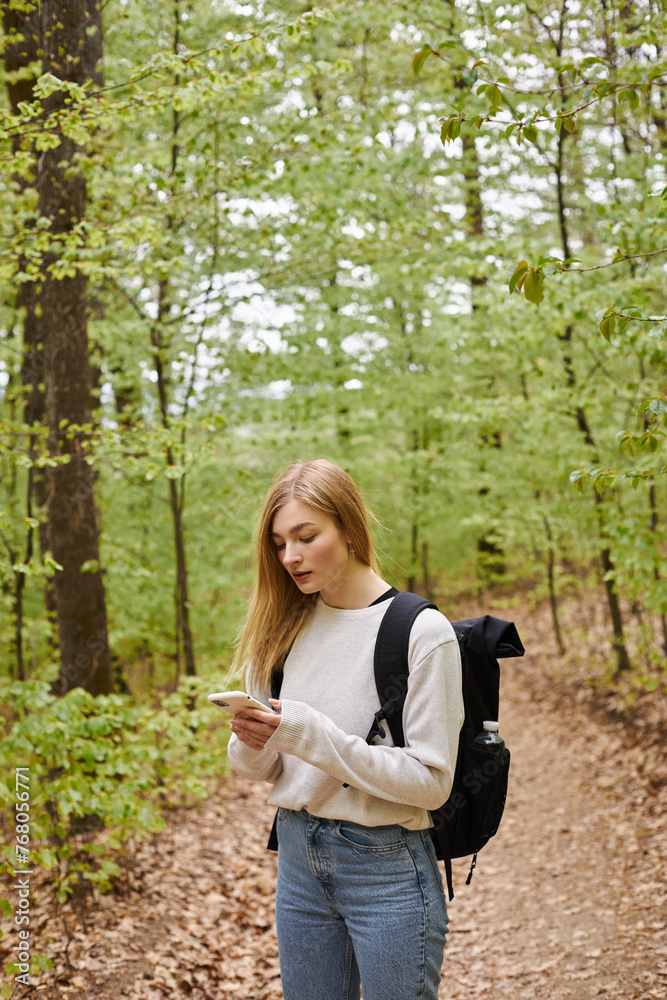 Young blonde female traveler with backpack holding phone hiking in forest and standing on path Young blonde female traveler with backpack holding phone hiking in forest and standing on path