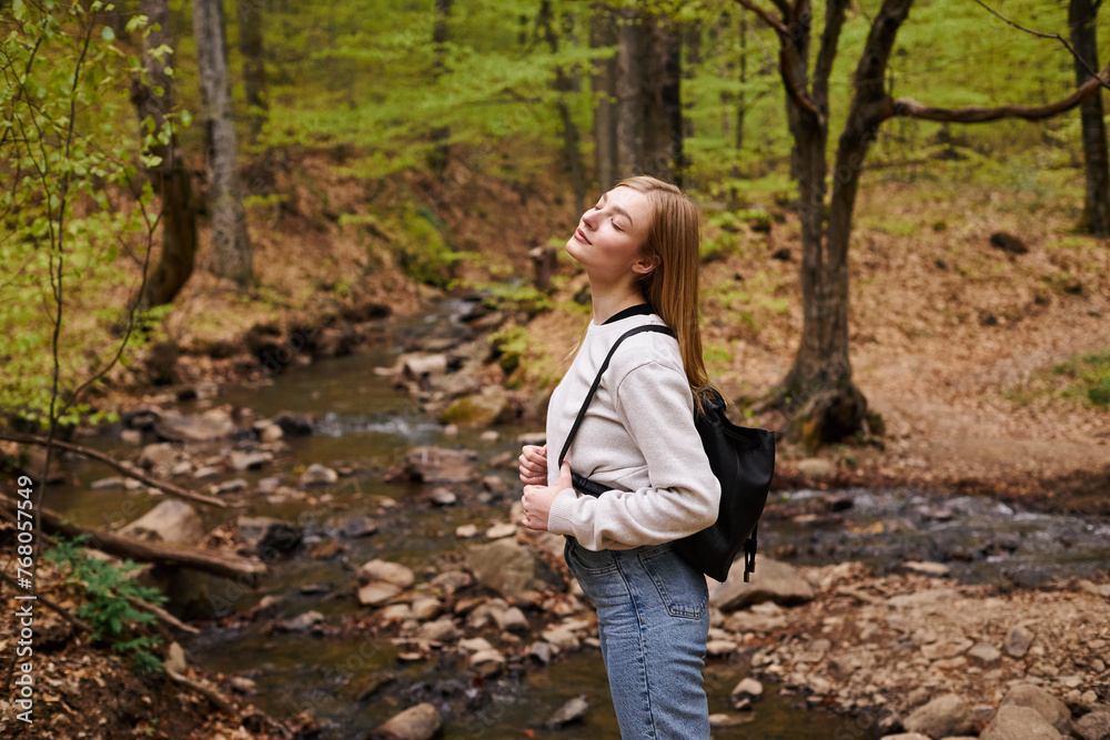 Young blonde female hiker standing with eyes closed in woods. Portrait of relaxed woman in forest