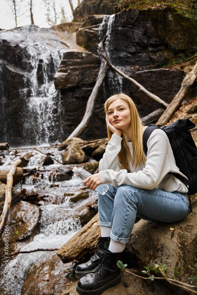 Smiling woman sitting with bottle of water and looking away in forest near waterfall Smiling woman sitting with bottle of water and looking away in forest near waterfall