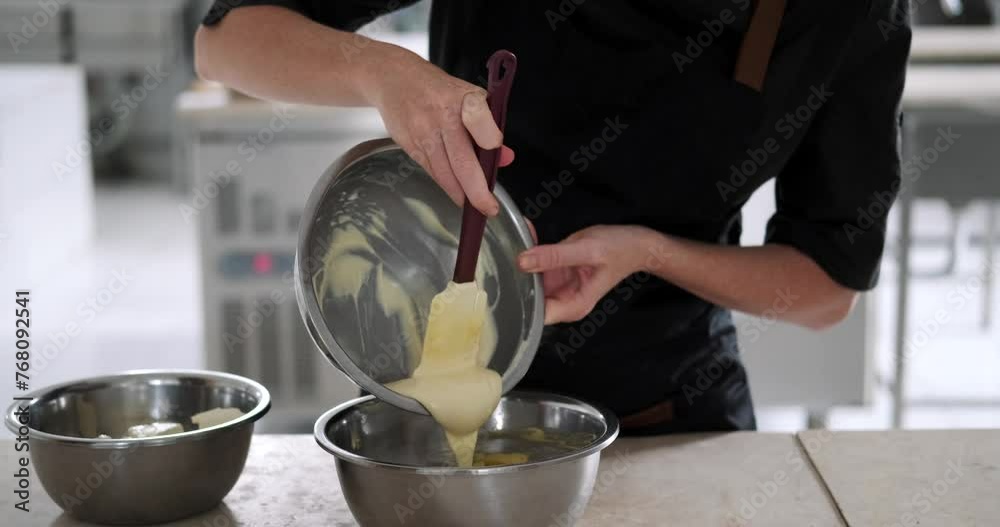Medium shot of the hands of a young woman pastry chef preparing buttercream for a cake and mixing two parts of the cream together in an iron bowl. The cooking process at a confectionery factory.