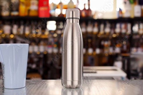 One metal vacuum-insulated bottle for water or coffee and a cup of coffee on a table on a bar background.