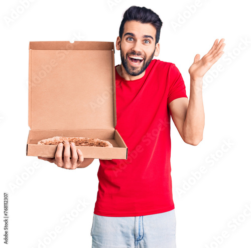 Young handsome man with beard holding delivery cardoboard with italian pizza celebrating victory with happy smile and winner expression with raised hands photo