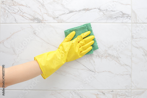crop of female hand in yellow rubber glove cleaning a white marble ceramic tiles on the wall in the bathroom. Spring general or regular cleaning concept.