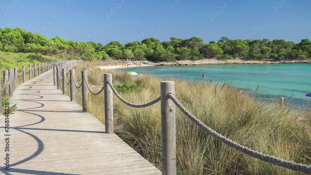Wooden path along Son Saura sandy beach with turquoise water surrounded by green pine trees in Menorca island, Spain