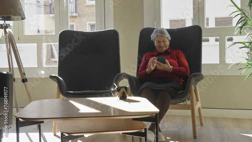 Single old woman sits on armchair and watching funny memories on mobile phone