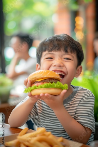 Excited happy asian little boy eating big burger at home