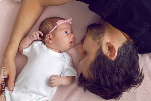 Close-up fashionable portrait of a man with hipster beard with baby girl lying on the bed in front of a window. Girl has a pink bow on her head and white clothes.