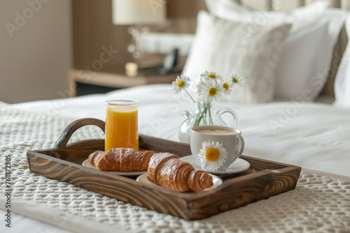 Bandeja de madera  conteniendo desayuno con taza de café, zumo de naranja y cruasanes y flores, sobre una cama 
 con ropa de cama blanca y fondo de habitación desenfocado