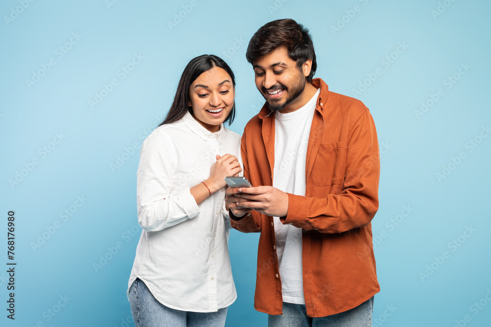 Excited indian couple looking at mobile phone using application, studio - obrazy, fototapety, plakaty 