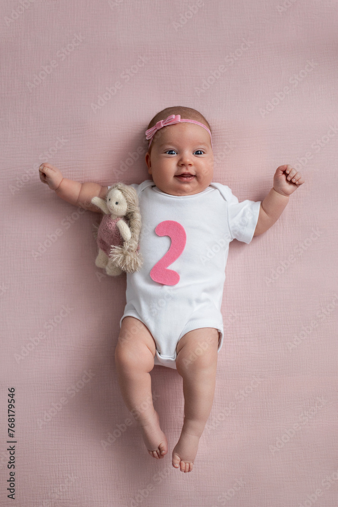 girl baby infant with a bow lies on the bed in a white bodysuit with a doll toy next to it, the child has a pink number 2 months on a pink background