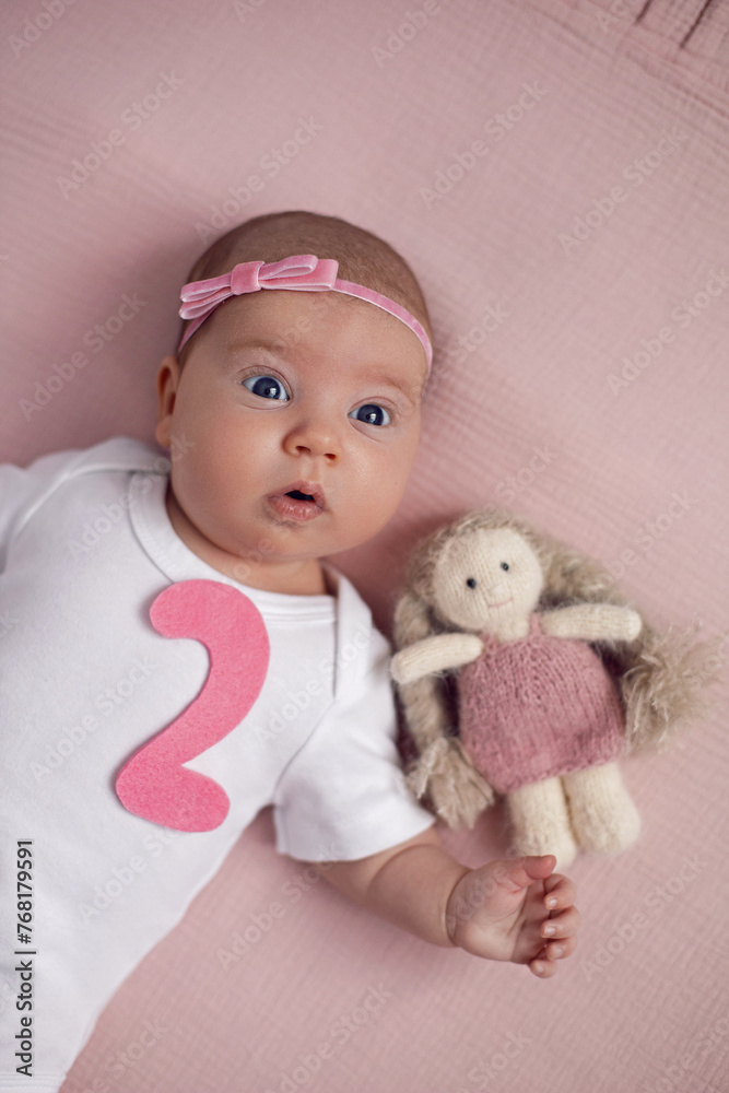 girl baby infant with a bow lies on the bed in a white bodysuit with a doll toy next to it, the child has a pink number 2 months on a pink background