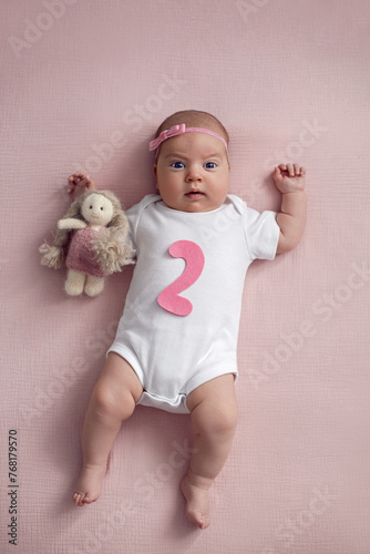 girl baby infant with a bow lies on the bed in a white bodysuit with a doll toy next to it, the child has a pink number 2 months on a pink background