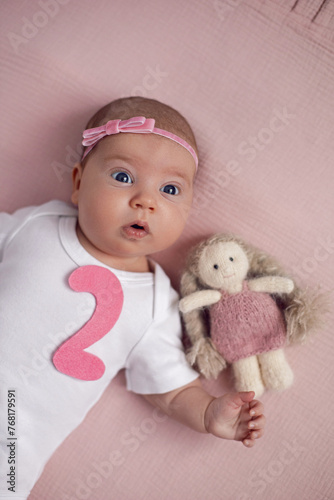 Obraz na płótnie girl baby infant with a bow lies on the bed in a white bodysuit with a doll toy next to it, the child has a pink number 2 months on a pink background w salonie