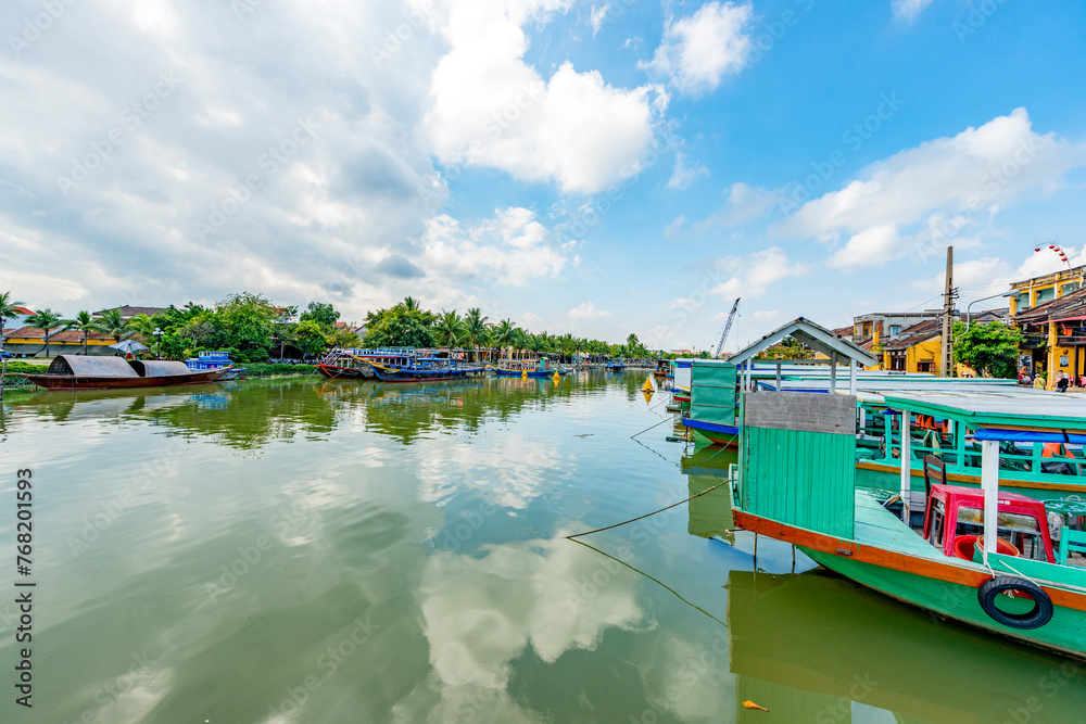 Obraz premium Wooden boats on the Thu Bon River in Hoi An Ancient Town (Hoian), Vietnam. Yellow old houses on waterfront reflected in river. Riverside Scenes in the Historic District of Hoi An, Vietnam