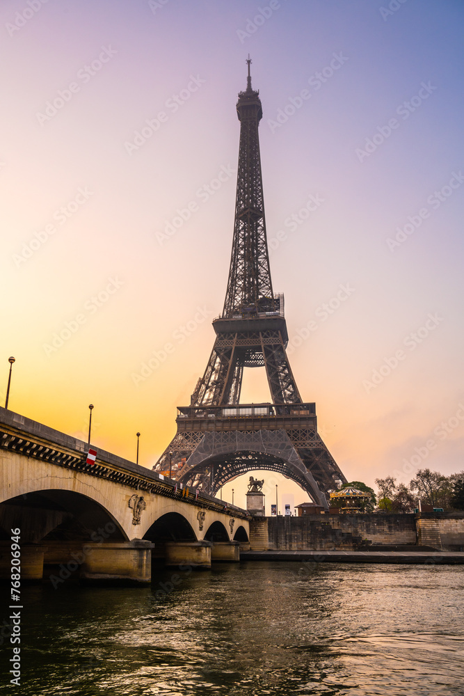 A captivating view of the Eiffel Tower and a bridge over the Seine River at sunrise. The sky is painted with warm hues, casting a serene atmosphere over Paris, France.