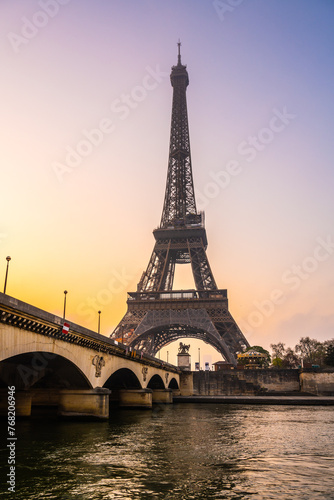 A captivating view of the Eiffel Tower and a bridge over the Seine River at sunrise. The sky is painted with warm hues, casting a serene atmosphere over Paris, France. © pyty