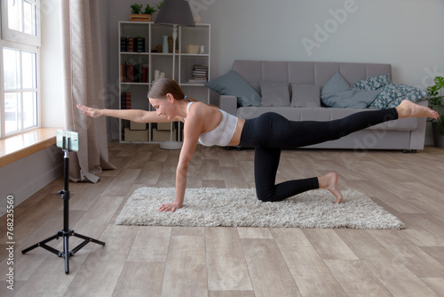 woman in black leggings and a white top does yoga at home on the carpet, recording an online fitness video for her audience with a smartphone standing on a tripod