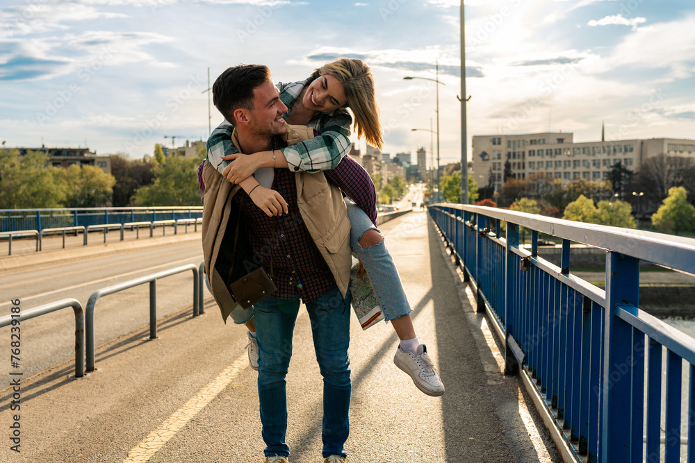 Strong handsome smiling man is holding piggyback his girlfriend on a bridge, they are having fun