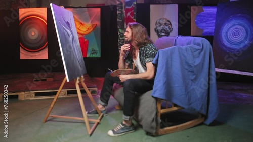 Side view of original painter sitting on wooden rocking chair covered with blue cloth inside art studio. Pensive caucasian artist examining masterpiece and prouding of work indoors. photo