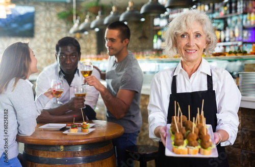 Smiling mature female waiter holding tray with pinchos at restaurant