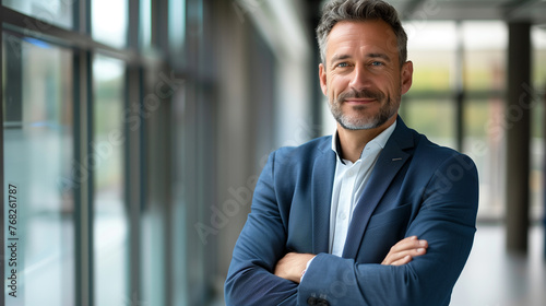 Portrait of mature businessman standing with arms crossed in office. Mature man in formalwear looking at camera and smiling. Business concept