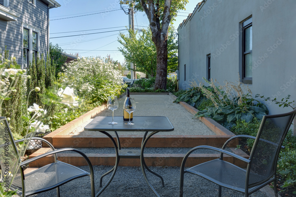 Naklejka premium A charming patio setup: Two chairs beside a table holding a wine bottle and glasses, set amidst a lush garden outside a beautiful traditional cottage.