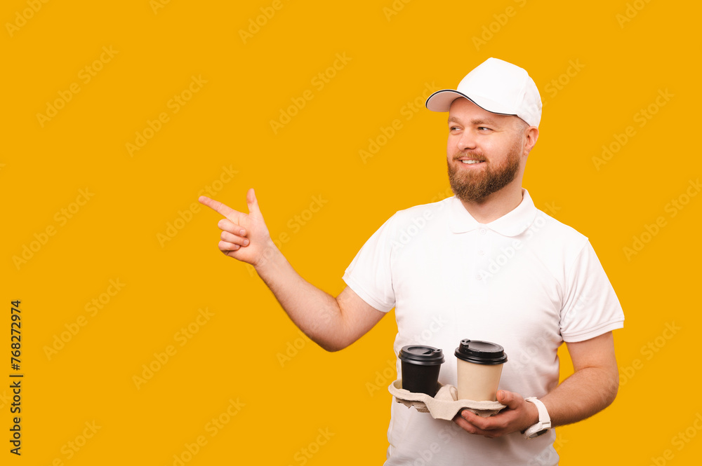 Smiling bearded delivery man is holding two take away cups and pointing aside at copy space.